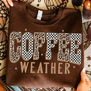 May include: A brown sweatshirt with the words "COFFEE WEATHER" in a bold, decorative font. The word "COFFEE" has a leopard print design and a black and white checkered pattern. The word "WEATHER" is in a simple, tan font.