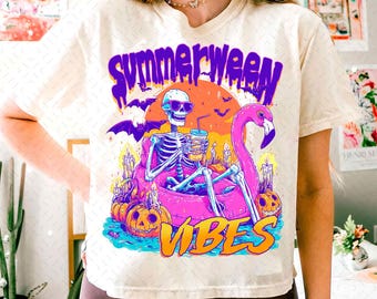 May include: Cream-colored t-shirt with a vibrant graphic. The design features a skeleton in sunglasses, holding a drink, floating on a pink flamingo pool float. The words "SUMMERWEEN VIBES" are displayed in purple and orange.