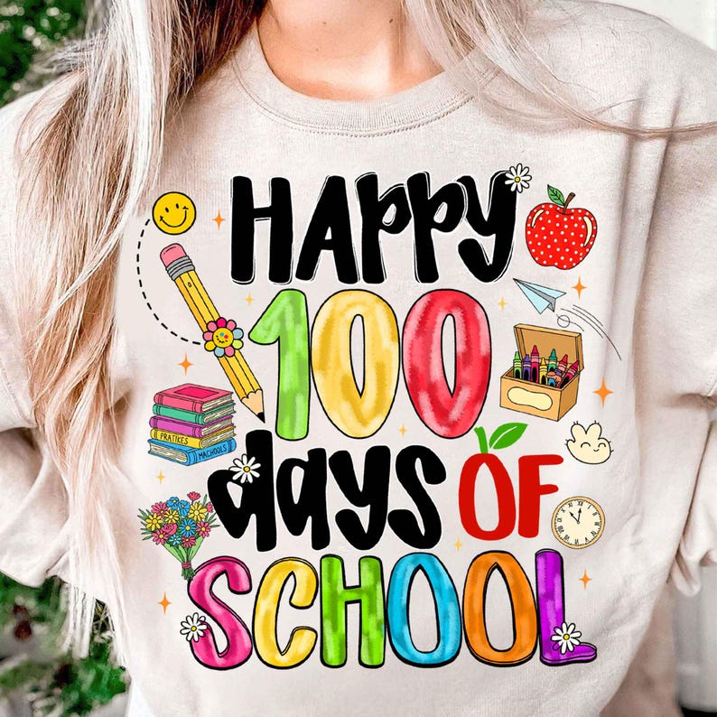 May include: Beige sweatshirt with a colorful graphic celebrating 100 days of school. The design includes the words "Happy 100 Days of School" with school-themed illustrations like a pencil, books, and an apple.