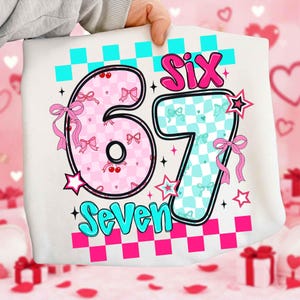May include: A white garment featuring the numbers "67" in a playful design. The number "6" is pink with bows and cherries, while the "7" is light blue with bows. The word "Six" is in pink, and "Seven" is in turquoise.