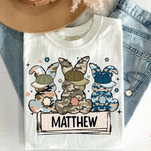Puede incluir: Camiseta blanca con un dise&ntilde;o de tres conejos de camuflaje con gorras de b&eacute;isbol. Los conejos est&aacute;n en fila y el texto "MATTHEW" est&aacute; debajo.
