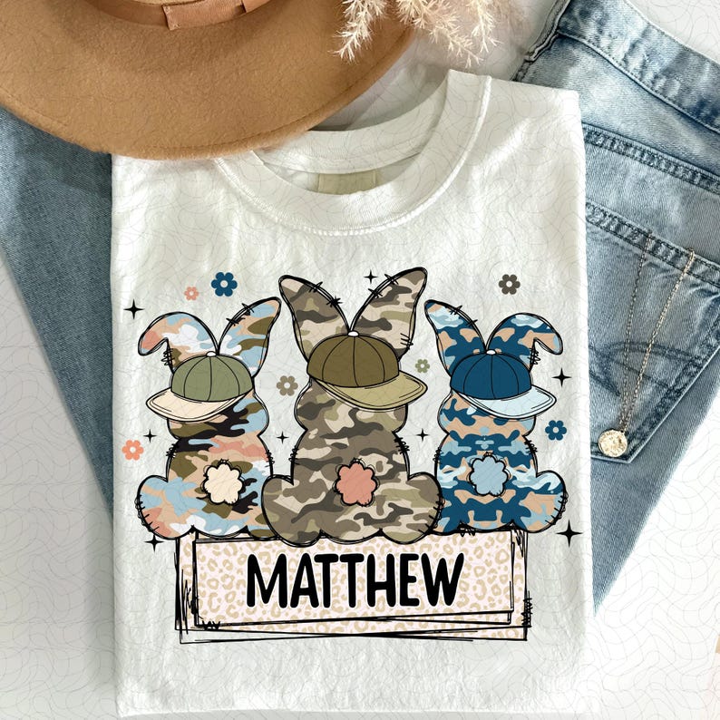 Puede incluir: Camiseta blanca con un dise&ntilde;o de tres conejos de camuflaje con gorras de b&eacute;isbol. Los conejos est&aacute;n en fila y el texto "MATTHEW" est&aacute; debajo.