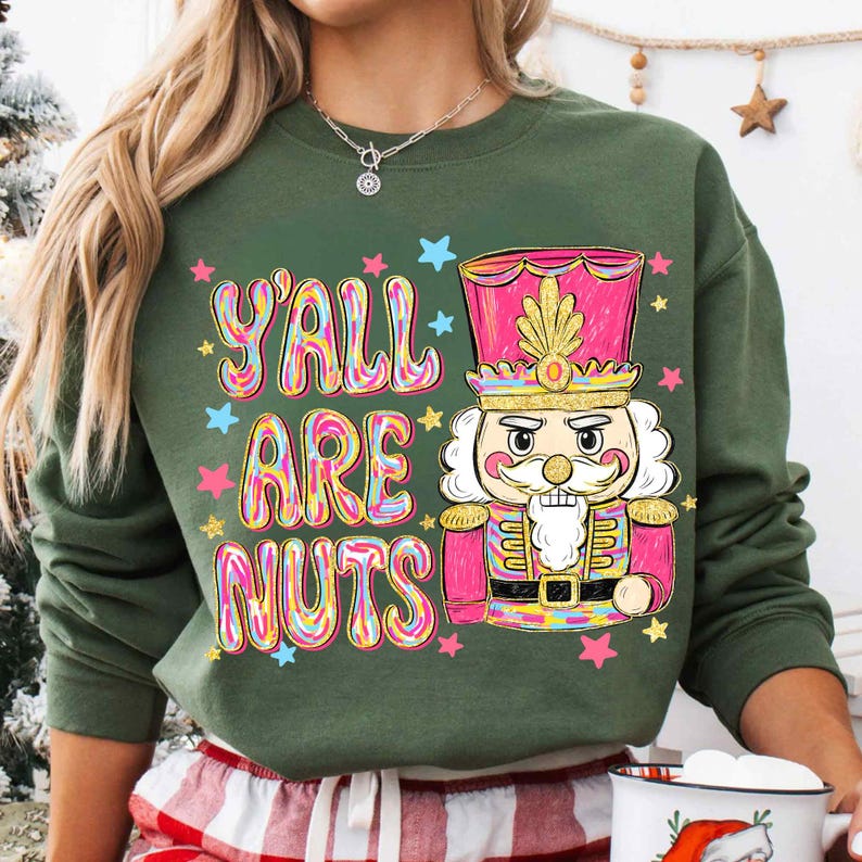 May include: Green sweatshirt with the text "Y'all Are Nuts" in colorful bubble letters, accompanied by a pink and gold nutcracker illustration and star accents. The sweatshirt is a festive holiday design.