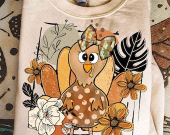 Peut inclure: Un sweat-shirt beige avec un motif de dinde de dessin animé. La dinde porte un nœud à carreaux et est entourée de fleurs orange et blanches et d'accents de feuilles noires. Le corps de la dinde a un motif à pois, sur fond neutre.