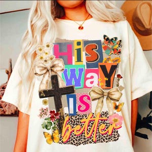 May include: Cream-colored t-shirt with the text "His Way Is Better" in a collage style. The design includes a cross, butterflies, flowers, and bows. The letters are in various colors and fonts.