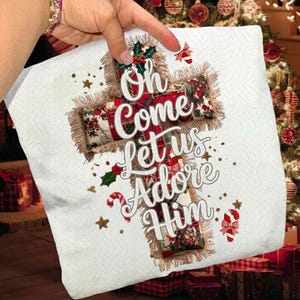 Puede incluir: Una almohada blanca con un diseño de cruz navideña. La cruz está decorada con patrones florales rojos y verdes y el texto "Oh Come Let Us Adore Him". La almohada tiene una textura similar a la arpillera y está adornada con bastones de caramelo y estrellas.