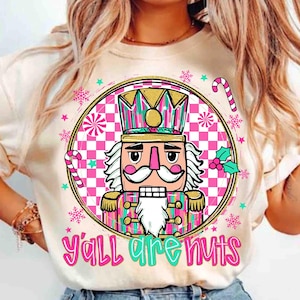 May include: A cream-coloured t-shirt with a festive nutcracker design. The image features a detailed nutcracker with a white beard and a crown, set within a circular frame. The text "Y'all are nuts" is displayed below, with candy canes and snowflakes.