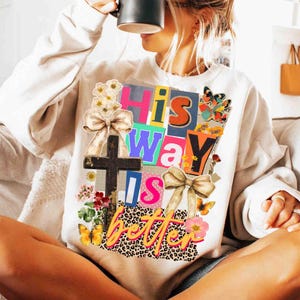 May include: Cream-colored sweatshirt with a colorful graphic that reads "His Way Is Better." The design includes a cross, bows, butterflies, and floral accents. The text is in various colors and fonts, with a leopard print background.