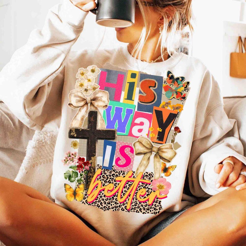May include: Cream-colored sweatshirt with a colorful graphic that reads "His Way Is Better." The design includes a cross, bows, butterflies, and floral accents. The text is in various colors and fonts, with a leopard print background.