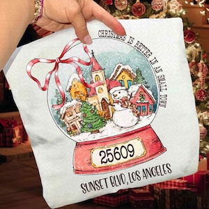 May include: A white pillow with a snow globe design. The snow globe features a snowman, houses, and a church in a winter scene. The text "Christmas is better in a small town" and "Sunset Blvd, Los Angeles" are visible. The pillow has a red bow.