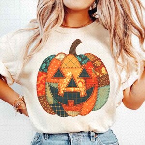 May include: A cream-coloured t-shirt with a patchwork pumpkin design. The pumpkin has a smiling jack-o'-lantern face with triangle eyes and a stitched mouth. The pumpkin is made of various fabric patterns in orange, green, and brown.
