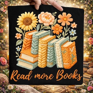 Może przedstawiać: Czarna poduszka z kwiatowym i książkowym wzorem. Wzór przedstawia słonecznik, pomarańczowe i brzoskwiniowe kwiaty oraz stos książek w odcieniach pomarańczu, żółci i niebieskiego. Tekst "Read more Books" jest napisany pomarańczowym skryptem.