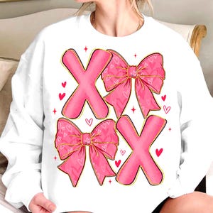 May include: White sweatshirt featuring pink X's and bows with heart patterns. The X's and bows have gold outlines, and small red hearts are scattered around the design. A Valentine's Day themed graphic.