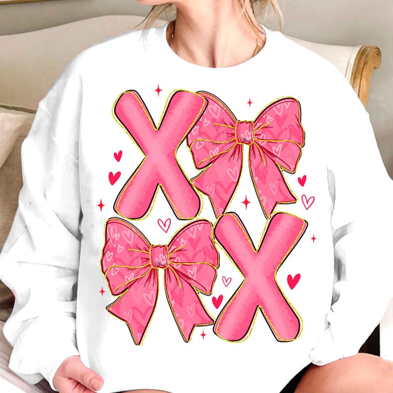 May include: White sweatshirt featuring pink X's and bows with heart patterns. The X's and bows have gold outlines, and small red hearts are scattered around the design. A Valentine's Day themed graphic.