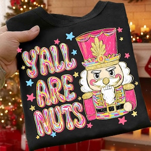 May include: Black t-shirt featuring the phrase "Y'all Are Nuts" in colourful bubble letters. The design includes a cartoon nutcracker in pink and gold, with stars. A festive holiday-themed graphic.