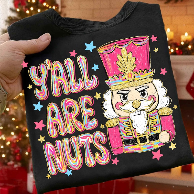 May include: Black t-shirt with the text "Y'all Are Nuts" in colorful bubble letters. A cartoon nutcracker in a pink and gold uniform is featured on the shirt, surrounded by stars. A festive holiday design.
