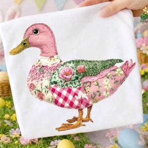 May include: A decorative duck applique on a white background. The duck features a pink head and beak, with a patchwork body of floral and gingham patterns in pinks and greens. The duck's feet are a golden brown color.
