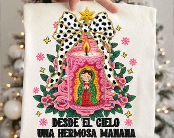 May include: A cream-colored t-shirt featuring a pink candle with a flame, a bow with black and white polka dots, and a floral arrangement. The shirt has the text "DESDE EL CIELO UNA HERMOSA MANANA" in black.