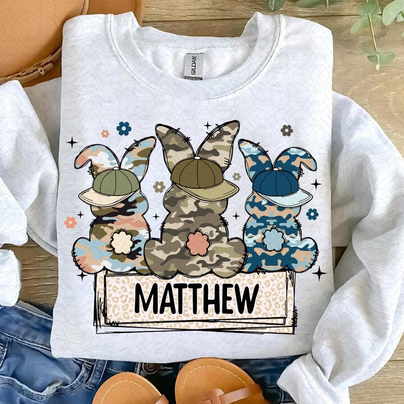 Puede incluir: Una sudadera blanca con un dise&ntilde;o de tres conejos de Pascua de camuflaje con sombreros. Los conejos est&aacute;n sentados en un letrero de madera con el nombre "MATTHEW" escrito en &eacute;l.