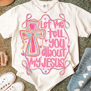 K&ouml;nnte beinhalten: Cremefarbenes T-Shirt mit einer bunten Grafik eines Kreuzes und dem pinkfarbenen Text "Let Me Tell You About My Jesus". Das Design enth&auml;lt rosa, blaue und gelbe Akzente. Eine silberne Halskette ist am Ausschnitt zu sehen.