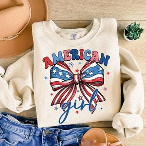 May include: Cream-colored sweatshirt with the words "AMERICAN girl" in red, blue, and pink lettering. A large bow graphic in red, white, and blue, with stars, is centered on the front. The sweatshirt is paired with denim jeans and sandals.
