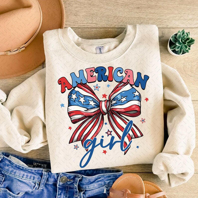 May include: Cream-colored sweatshirt with the words "AMERICAN girl" in red, blue, and pink lettering. A large bow graphic in red, white, and blue, with stars, is centered on the front. The sweatshirt is paired with denim jeans and sandals.