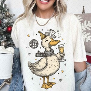 May include: Cream-colored t-shirt featuring a cartoon duck wearing a party hat and scarf, holding a champagne glass. The text "HAPPY NEW YEAR" is above the duck. A disco ball and stars are also in the design.