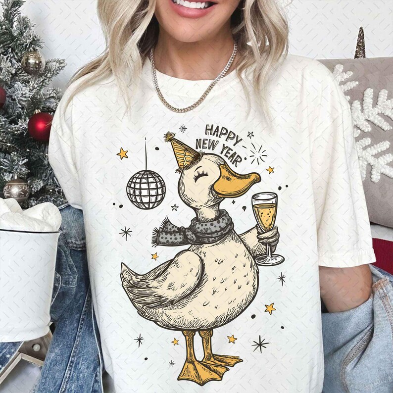 May include: Cream-colored t-shirt featuring a cartoon duck wearing a party hat and scarf, holding a champagne glass. The text "HAPPY NEW YEAR" is above the duck. A disco ball and stars are also in the design.