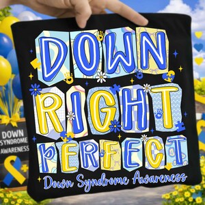 May include: A black t-shirt featuring the text DOWN RIGHT PERFECT in large, colorful, block-style letters with blue and yellow patterns. The design includes small icons of daisies, puzzle pieces, and handprints, with the text Down Syndrome Awareness below.