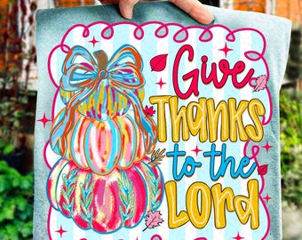 May include: A decorative square pillow featuring a stacked pumpkin design with a bow and the text "Give Thanks to the Lord." The pumpkins are in shades of pink, orange, and blue, set against a light blue striped background.