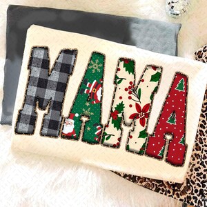Peut inclure: Un coussin crème avec le mot "MAMA" en grandes lettres décoratives. Les lettres sont remplies de motifs de Noël variés, dont du tartan, le Père Noël, du houx et des sapins. Les lettres sont bordées d'une couture marron.