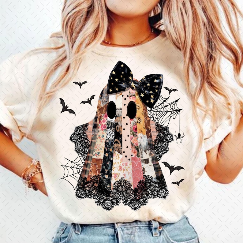 May include: Cream-colored t-shirt featuring a patchwork ghost design with a black bow adorned with gold stars. The ghost has black eyes and is decorated with floral and plaid patterns. Black lace trim, spiderwebs, spiders, and bats complete the Halloween theme.