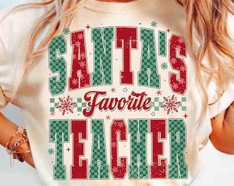 Puede incluir: Camiseta color crema con el texto "SANTA'S Favorite TEACHER" en letras de bloque rojas y verdes. El diseño incluye copos de nieve y estrellas, creando un tema festivo navideño. La camiseta tiene cuello redondo clásico y mangas cortas.