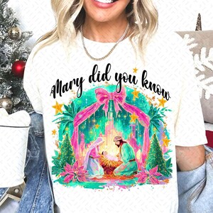 May include: White t-shirt with the text "Mary did you know" in black script. The shirt features a colorful graphic of the Nativity scene with pink ribbons, stars, and Christmas trees. The design is festive and holiday-themed.