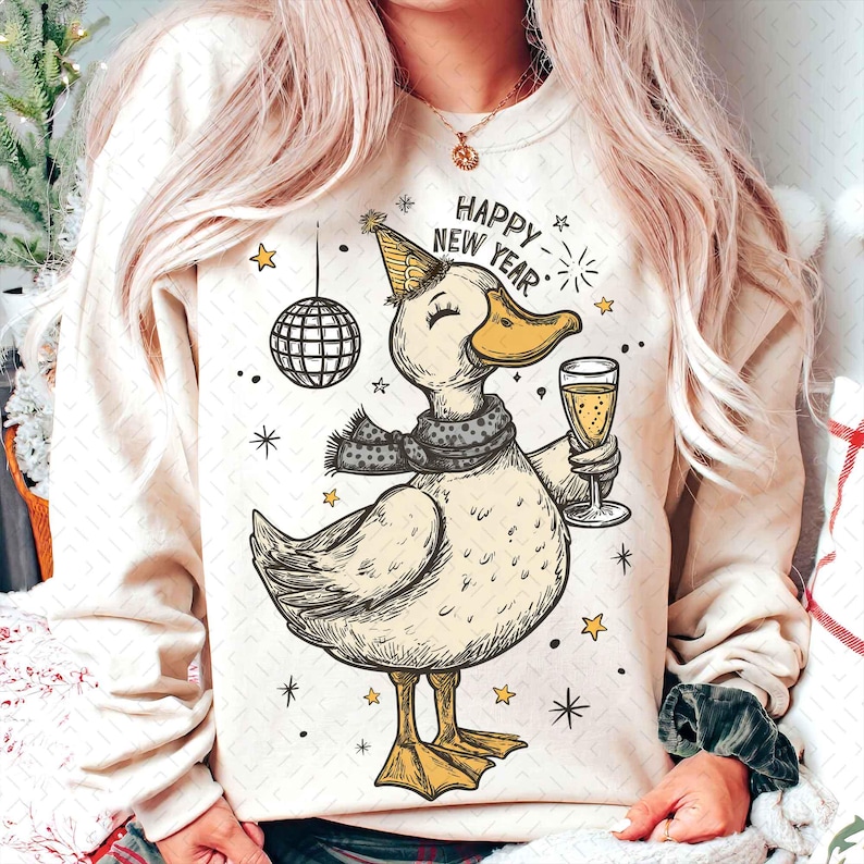 May include: Cream-colored sweatshirt featuring a cartoon duck wearing a party hat and scarf, holding a champagne glass. The duck is surrounded by stars and a disco ball, with the words "Happy New Year" above. A festive design for the holiday season.