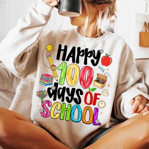 May include: Cream-colored sweatshirt with a colorful graphic that reads "Happy 100 Days of School." The design includes a smiling pencil, books, an apple, and a clock. The text is in various colors.