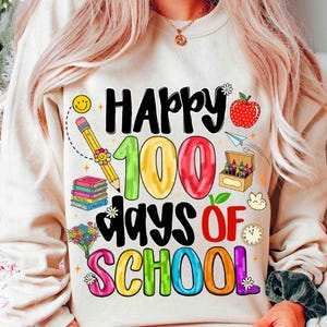 May include: Cream-colored sweatshirt with a colorful graphic celebrating "Happy 100 Days of School." The design includes a smiling pencil, books, an apple, and a box of crayons. The text is in various colors, including black, green, red, blue, and purple.