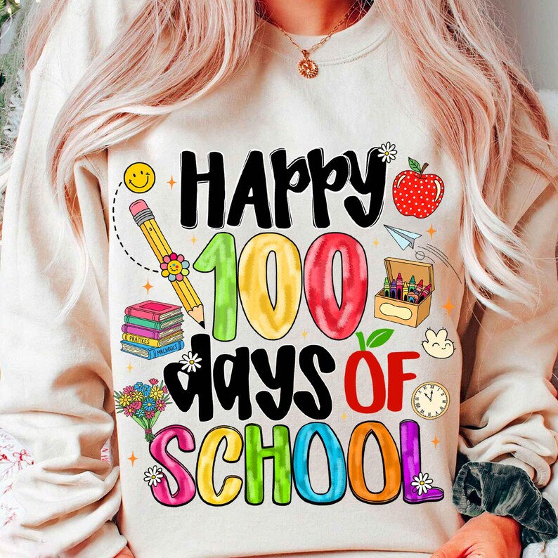 May include: Cream-colored sweatshirt with a colorful graphic celebrating "Happy 100 Days of School." The design includes a smiling pencil, books, an apple, and a box of crayons. The text is in various colors, including black, green, red, blue, and purple.
