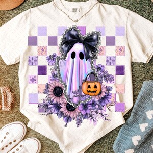 Könnte beinhalten: Weißes T-Shirt mit einem Halloween-Geister-Design. Der Geist ist lila und schillernd, mit einer schwarzen Schleife und einem Kürbis. Das Shirt hat ein Patchwork-Design in Lila- und Rosatönen mit Sonnenblumen und anderen Blumen.
