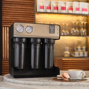 Puede incluir: Un sistema de filtración de agua de acero inoxidable con cuatro cartuchos de filtro. El sistema tiene una pantalla digital y dos manómetros. El sistema está sentado en una encimera de mármol junto a una taza de café y dos macarons.
