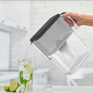 Puede incluir: Una jarra filtrante de agua gris y transparente vierte agua en un vaso lleno de hielo, rodajas de lima y menta. La jarra tiene una tapa negra y un asa blanca. El vaso está sobre una superficie blanca.