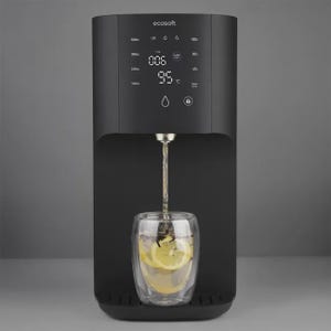 Puede incluir: Un dispensador de agua Ecosoft negro mate con una interfaz táctil digital que muestra 006 TDS y 95 grados Celsius. Un vaso transparente de doble pared con agua y rodajas de limón se sitúa bajo el surtidor, que está vertiendo agua caliente.