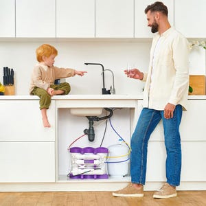 Puede incluir: Una escena de cocina con un sistema de filtración de agua. Un hombre sostiene un vaso de agua, mientras un niño señala un grifo negro. El sistema de filtración es blanco y morado, con varios tubos y un tanque blanco. La cocina tiene armarios blancos.