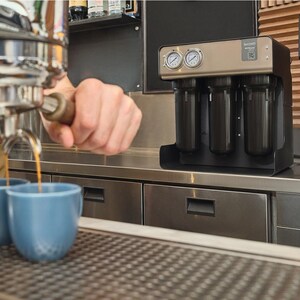 Puede incluir: Una máquina de espresso comercial con acabado de acero inoxidable dispensa café en dos tazas de cerámica azules. Un sistema de filtración de agua negro y dorado está sobre la encimera. La máquina está en un café.