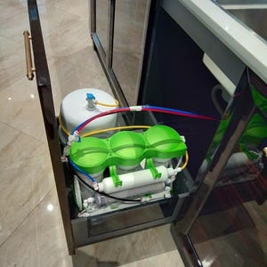 May include: A water filtration system installed under a kitchen sink. The system includes a white tank, three green filter canisters, and a black pump. The system is connected to plumbing with blue, red, and yellow tubing.