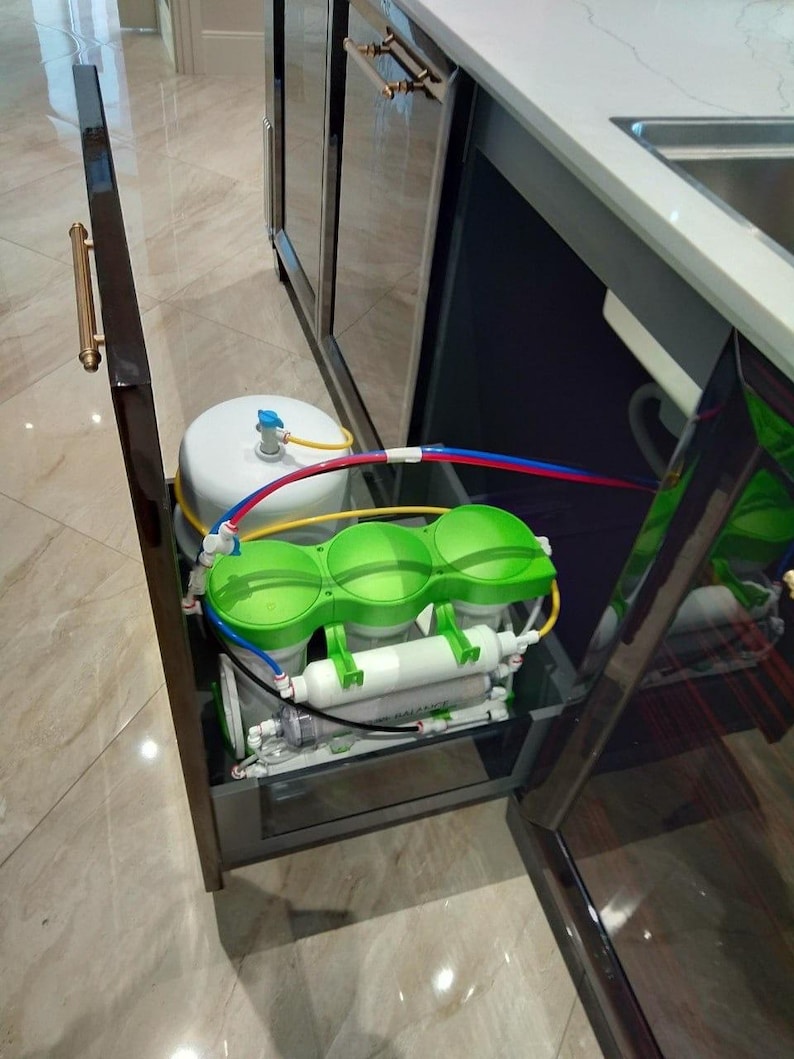 May include: A water filtration system installed under a kitchen sink. The system includes a white tank, three green filter canisters, and a black pump. The system is connected to plumbing with blue, red, and yellow tubing.