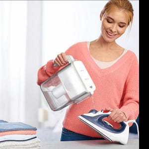 Puede incluir: Una mujer vierte agua de un recipiente blanco en una plancha azul y blanca. La plancha está sobre una mesa junto a una pila de ropa doblada. La mujer lleva un suéter rosa.