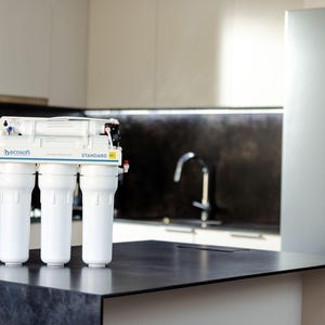 Puede incluir: Un sistema de filtración de agua blanco con cuatro filtros y el texto "by eco soft STANDARD" en la parte superior. El sistema está colocado sobre una encimera negra.