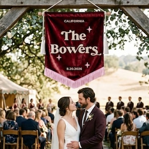 May include: A deep red velvet pennant banner featuring the white text CALIFORNIA, The Bowers, and 8.20.2026, accented with white star icons and a light pink fringe trim. The banner hangs from a wooden dowel with a white cord for display.