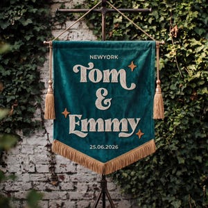 May include: A dark teal velvet pennant banner featuring embroidered cream-coloured text that reads NEWYORK, Tom & Emmy, and 25.06.2026. The banner is adorned with four small orange star icons, gold-coloured tassels on each side, and gold fringe at the bottom.